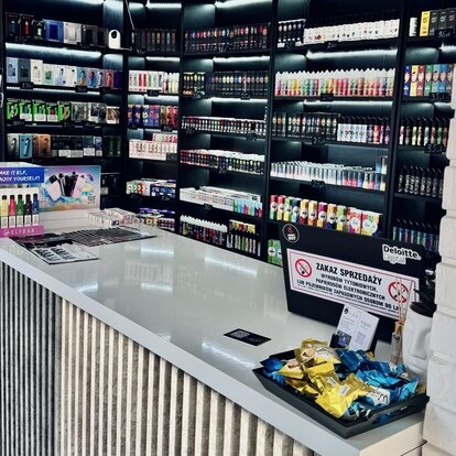 Wnętrze dobrze zaopatrzonego sklepu vape shop, z półkami pełnymi różnych e-papierosów i akcesoriów, oraz pracownikiem za ladą doradzającym klientowi.